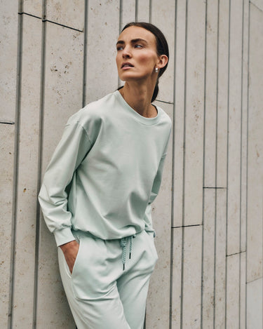 Woman wearing a light green sweatshirt and pants against a textured wall.